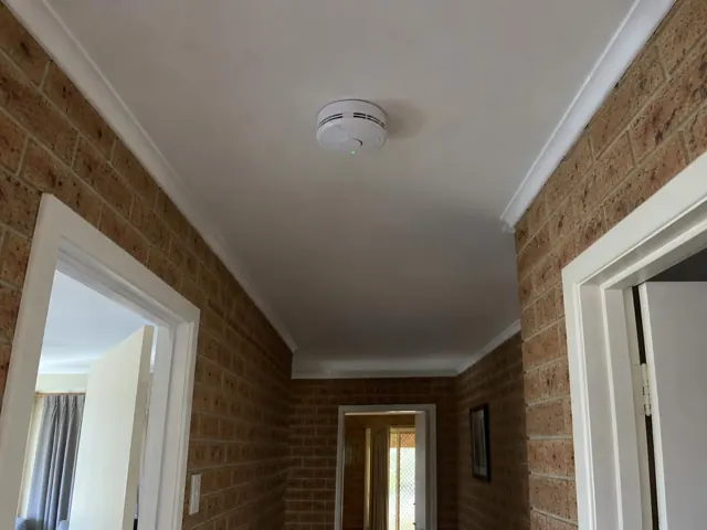 Ceiling-mounted smoke alarm in a brick hallway with white trim in Lake Munmorah